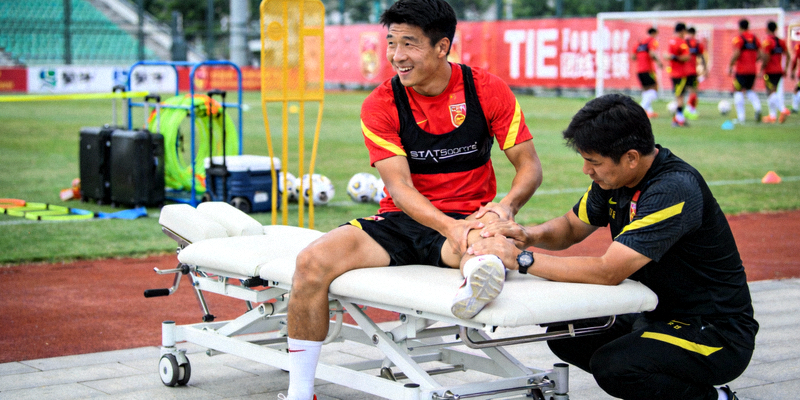 队医正在为一名国家队球员进行康复治疗，背景是训练场